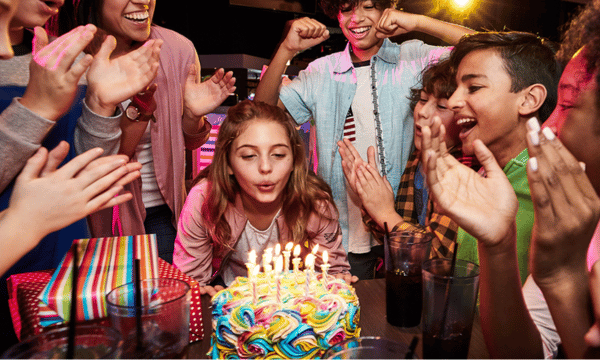 Girl blowing out candles on a colorful birthday cake surrounded by celebrating friends at Outta Boundz